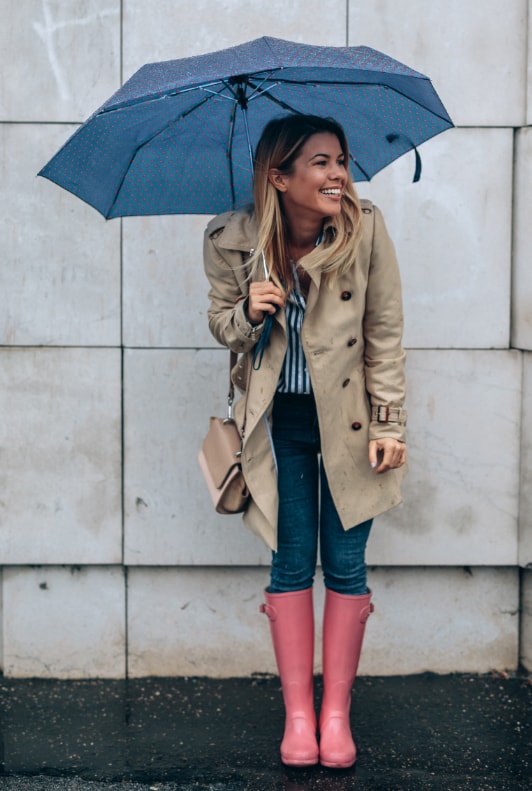 Blonde Frau mit Regenschirm, Trenchcoat und pinken Gummistiefeln