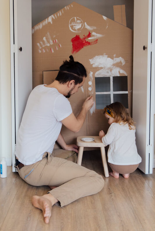 Vater und Kind bauen zusammen ein Haus aus Pappe und bemalen es mit buter Farbe