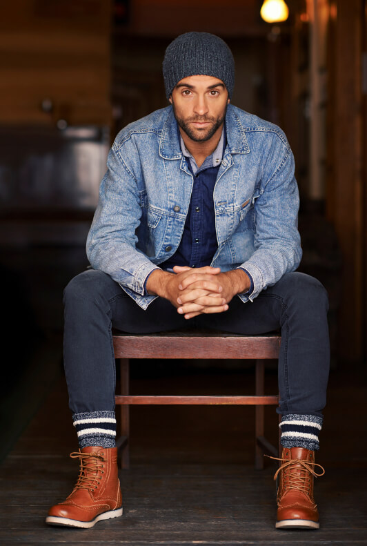 Mann mit Mütze auf Stuhl sitzend in blauer Jeansjacke und braunen Stiefeln