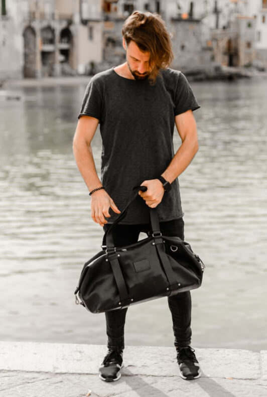 Junger Mann in schwarzem T-Shirt und schwarzer Hose trägt eine schwarze Umhängetasche aus Leder