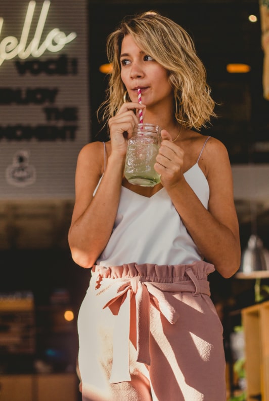 Junge Frau miit sommerlichen Papberbag-Hose in Rosa trinkt mit Strohalm einen Eistee